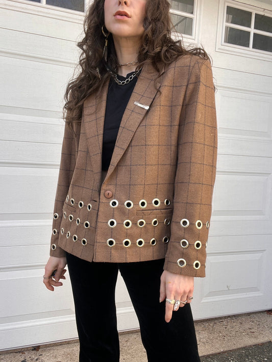 Woman wearing a brown checked blazer with circular embellishments, paired with a black top and pants, standing in front of a white garage door.