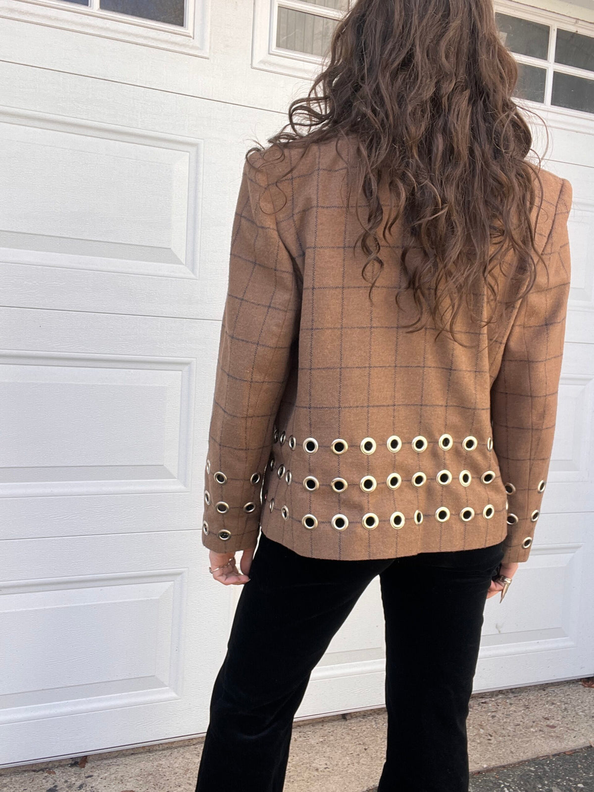 A person seen from behind wearing a checkered blazer with grommet details on the bottom standing in front of a white garage door.