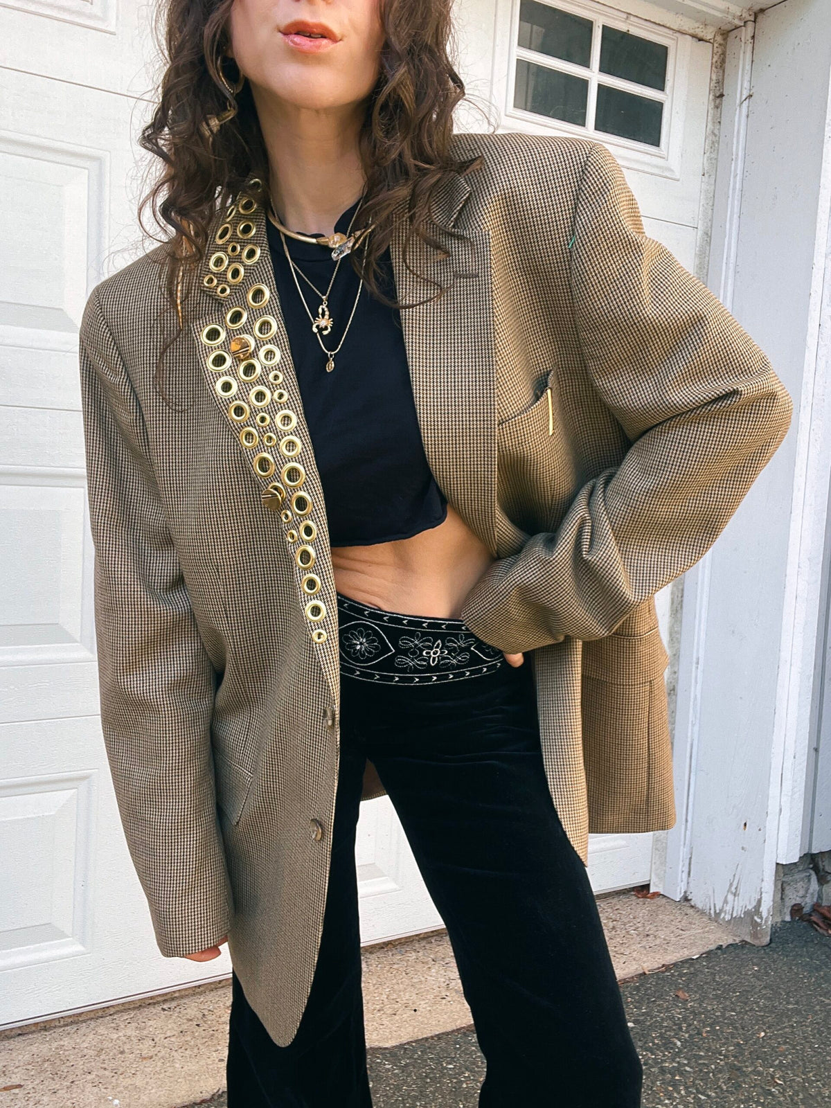 A person in a black crop top and high-waisted pants, wearing a patterned oversized blazer and layered necklaces.