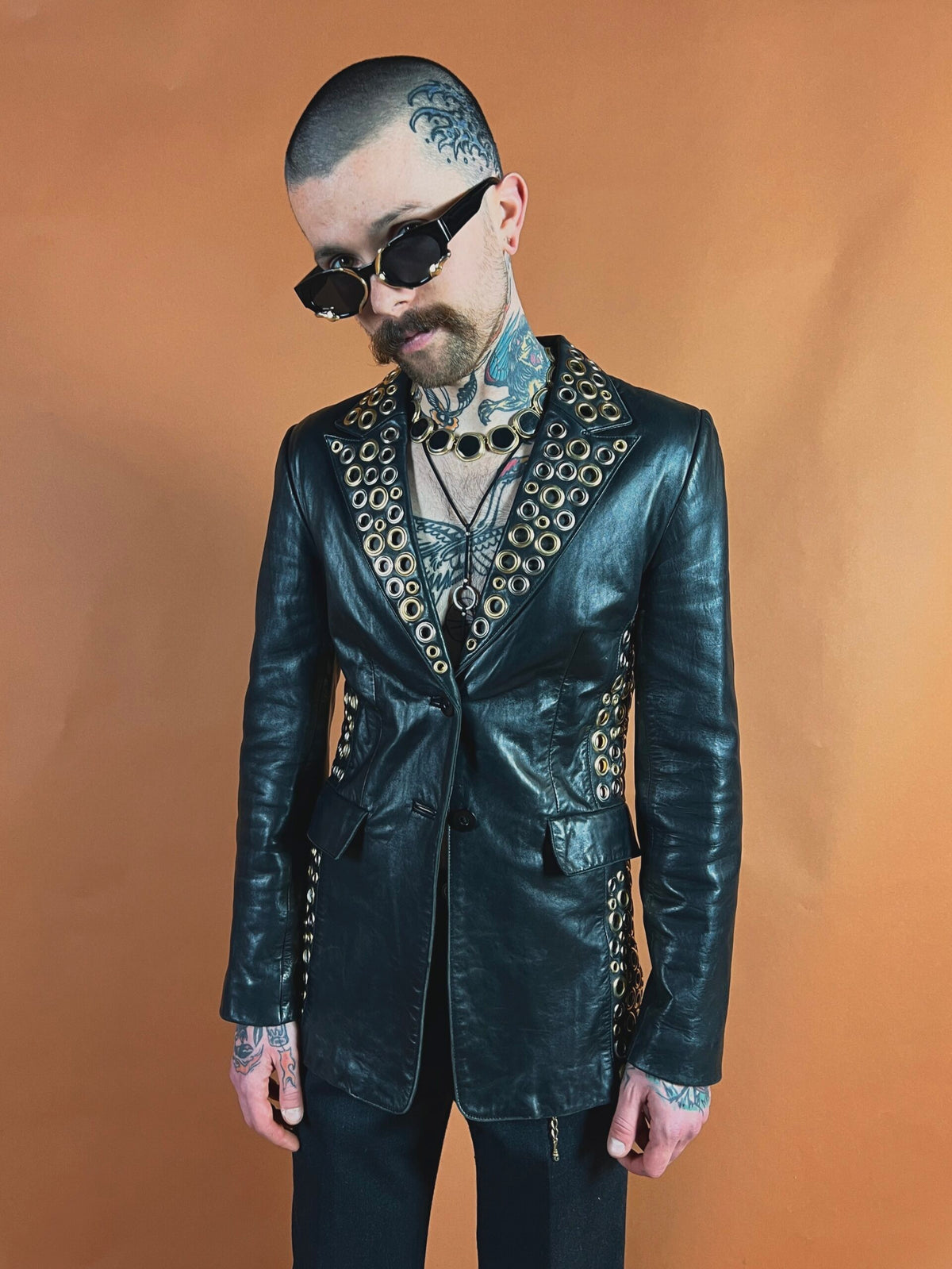Man with tattoos wearing sunglasses and a black leather jacket against an orange background.