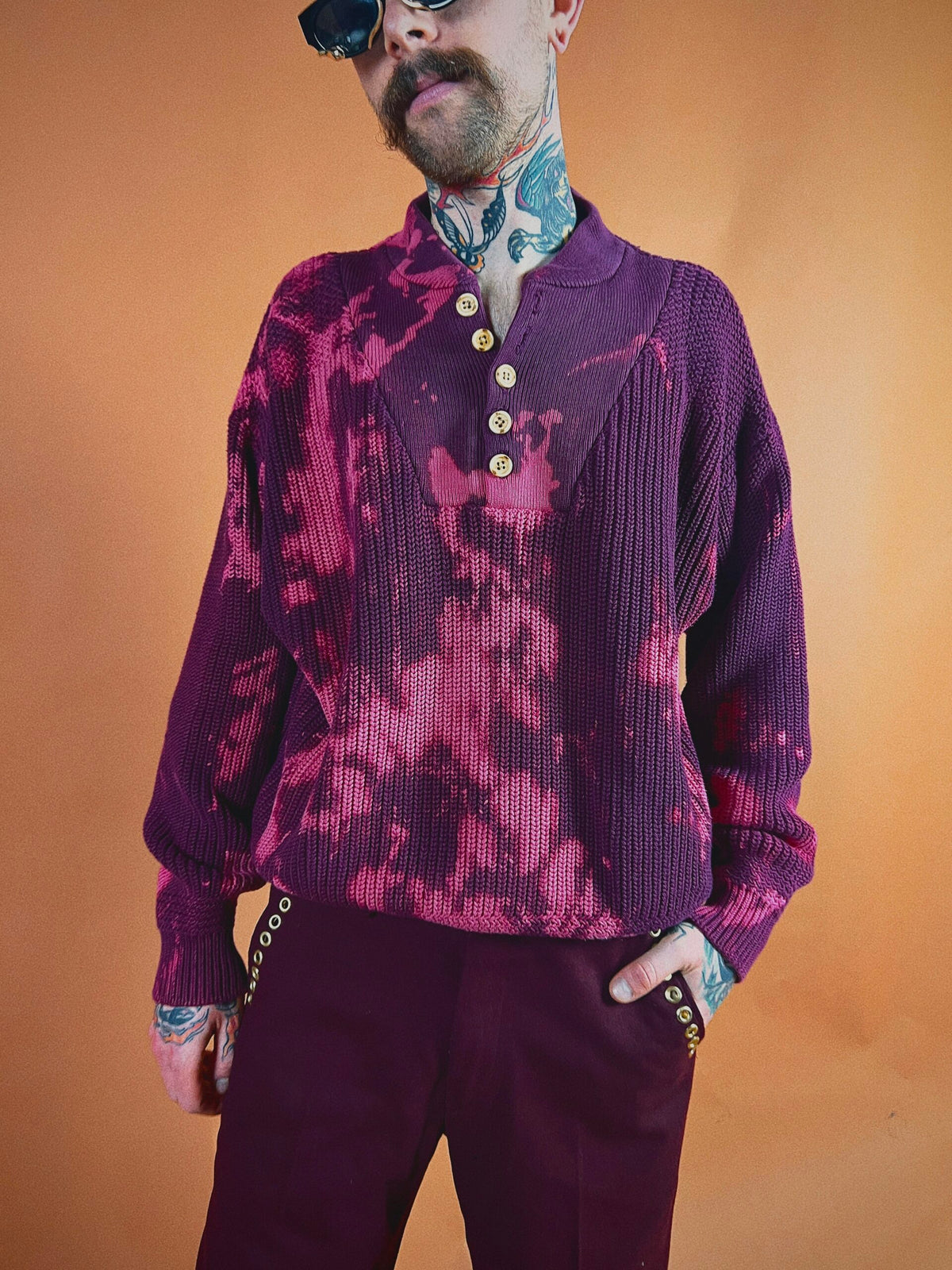 Man standing in a pose wearing a patterned purple shirt and maroon pants against an orange background.