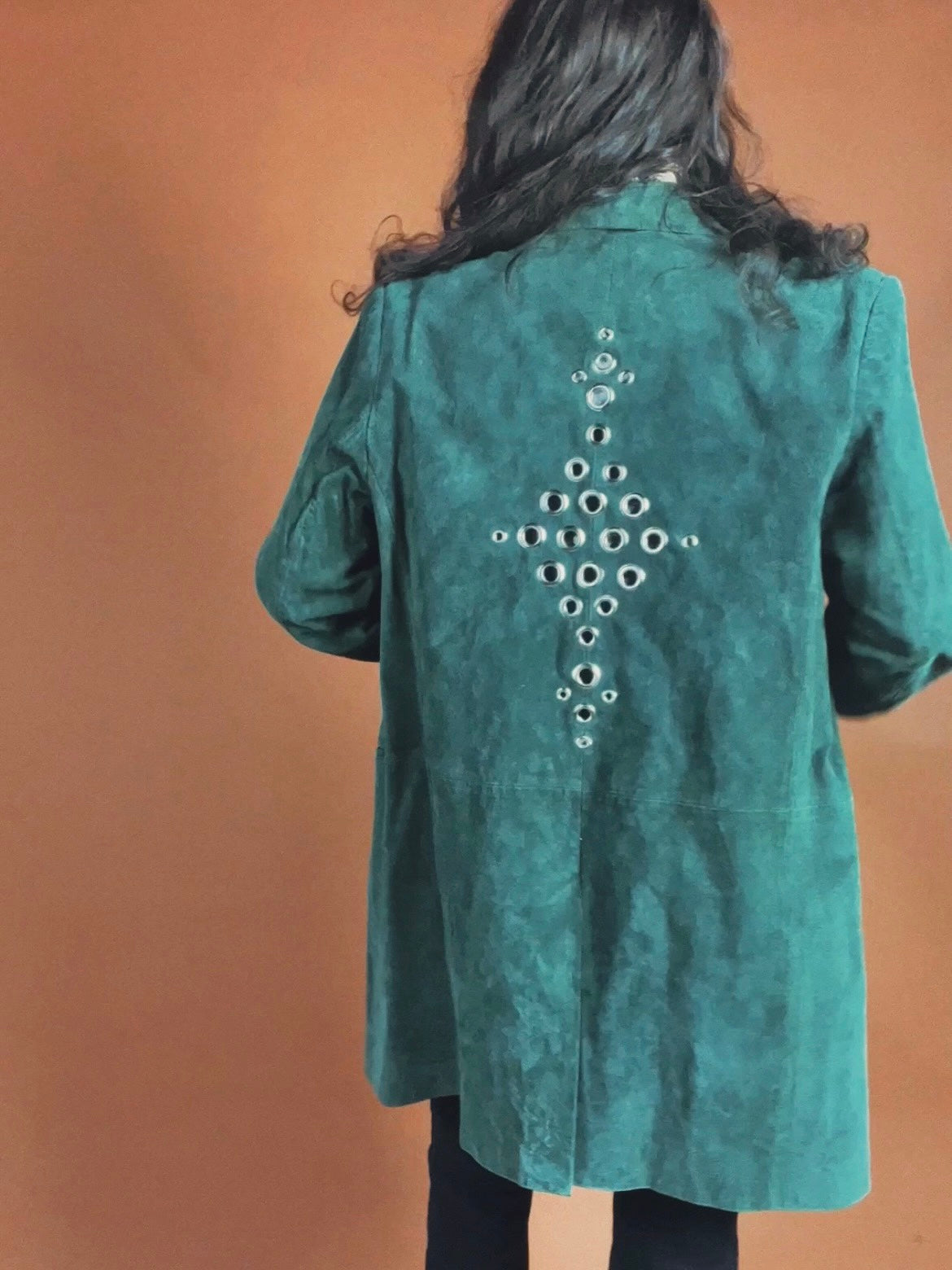 A person with long hair seen from behind wearing a teal suede jacket with a decorative pattern of metal eyelets on the back.