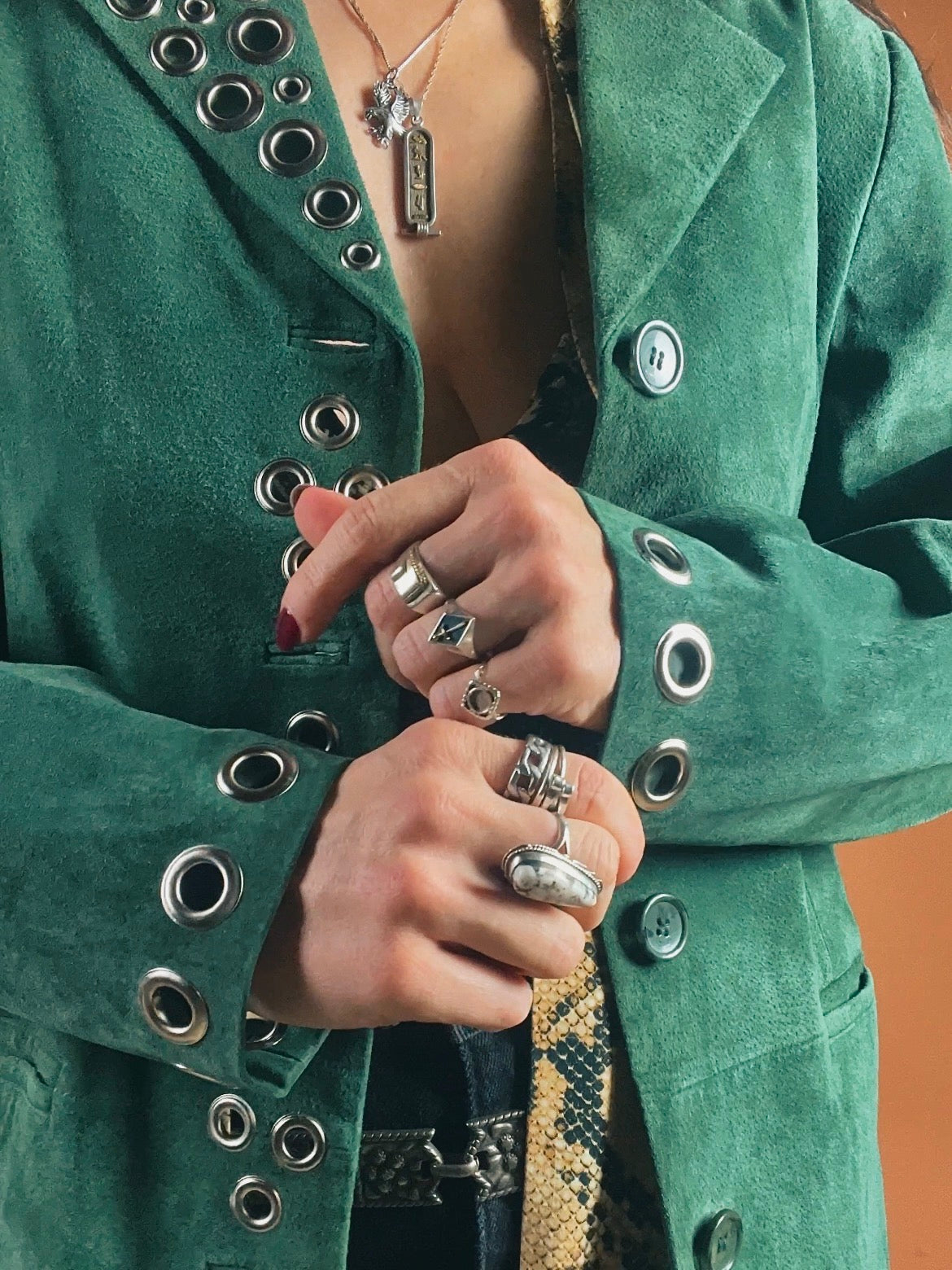 A person in a green jacket adorned with metal grommets, clutching the lapel with hands featuring multiple rings.