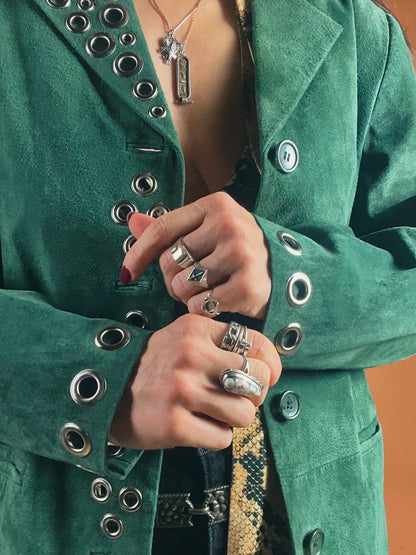 A person in a green jacket adorned with metal grommets, clutching the lapel with hands featuring multiple rings.