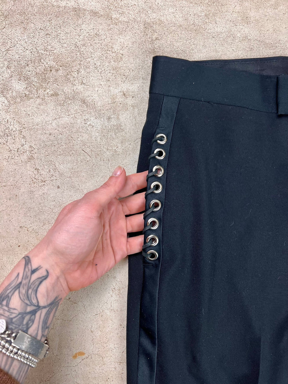 A hand with tattooed fingers holds open the side of a black garment featuring silver eyelet details.