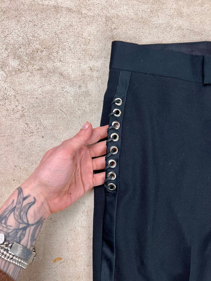 A hand with tattooed fingers holds open the side of a black garment featuring silver eyelet details.