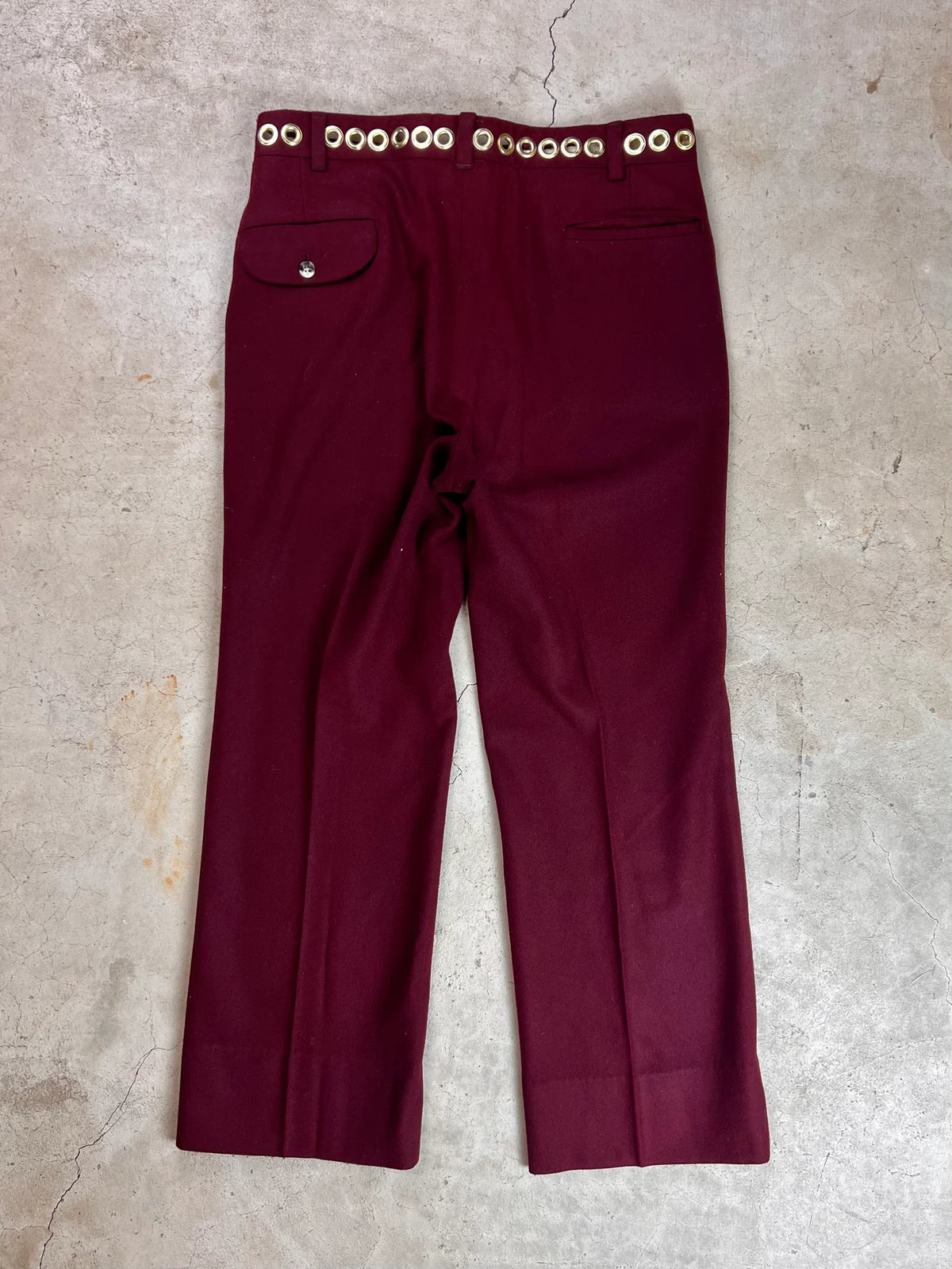 A pair of maroon pants with high waist and gold button details laid flat on a concrete surface.