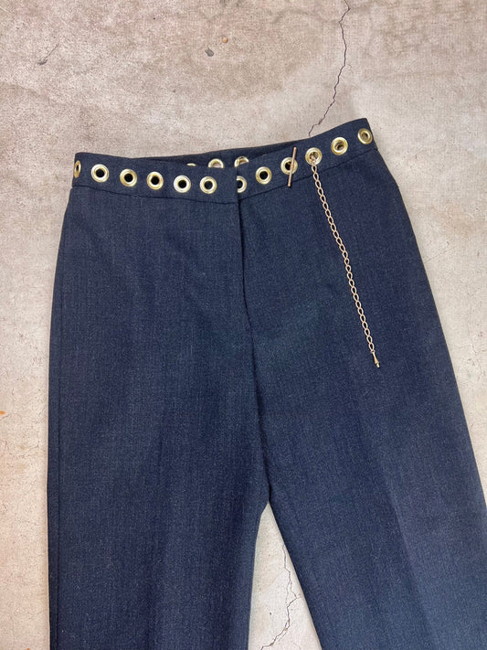 Dark blue trousers with eyelet belt loops and a chain detail on a concrete background.