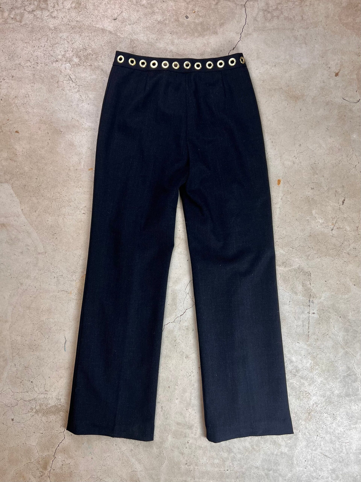Black trousers with a decorative grommet waistband on a concrete surface.
