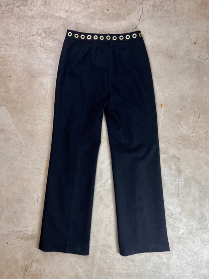 Black trousers with a decorative grommet waistband on a concrete surface.