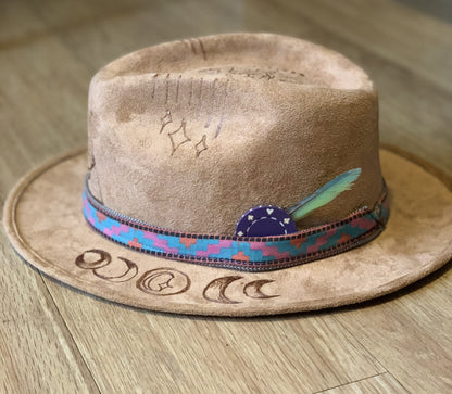 A suede hat wood burned with accessories with a feather on top of it.