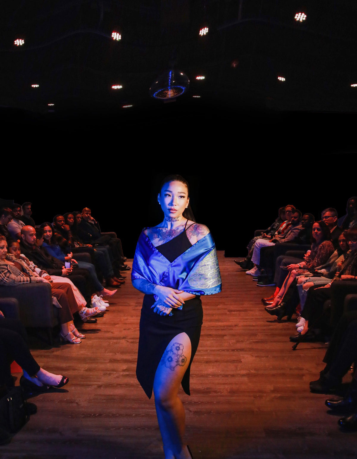 A woman standing on a runway in front of a crowd wearing a Hand-painted iridescent blue dragonfly wing scarf.
