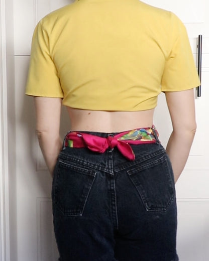 The back view of a woman wearing a yellow crop top and black jeans with a hand-painted belt.