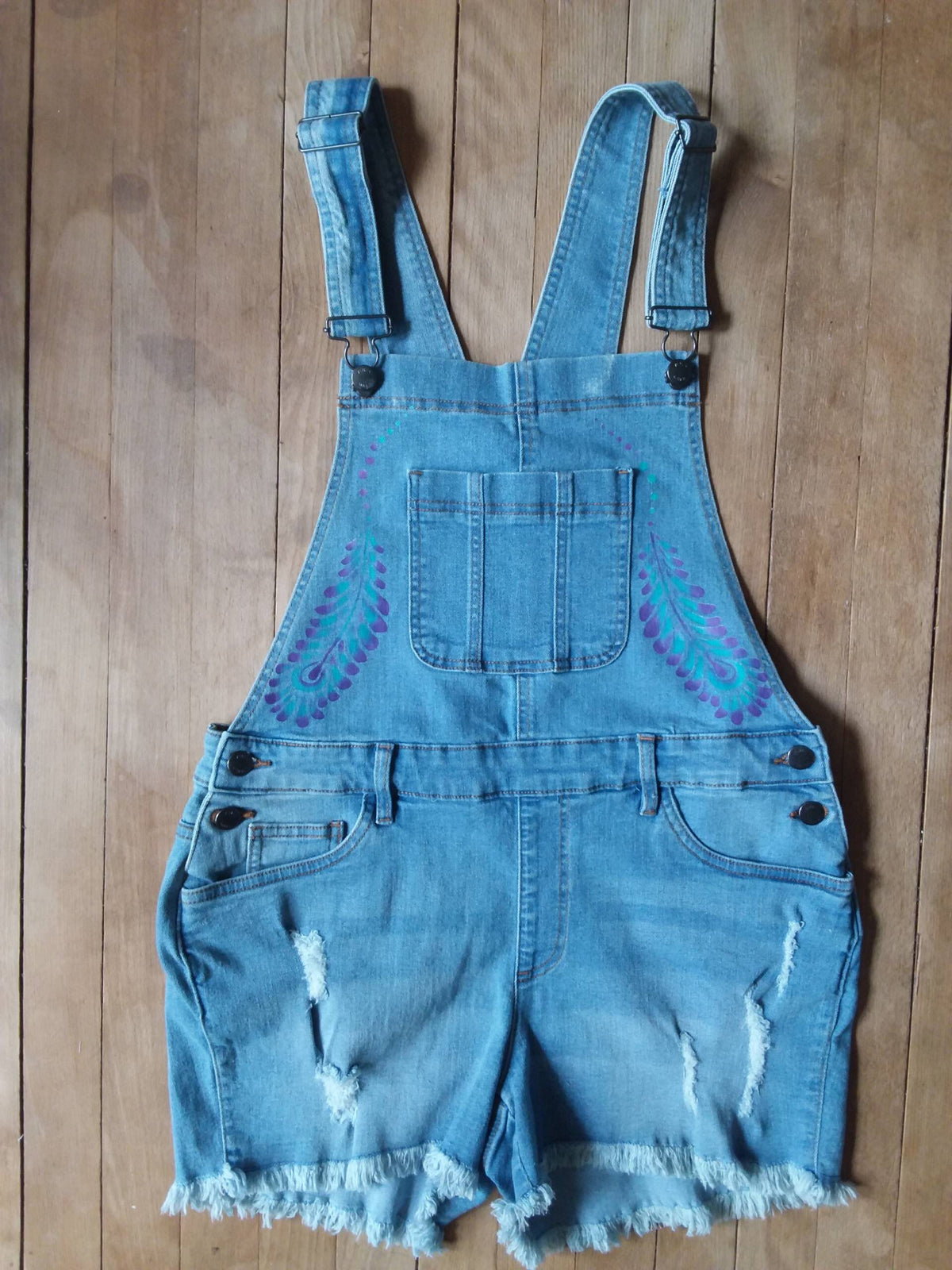Hand painted ladies denim overalls with distressed details have a lovely purple and teal feather design.
