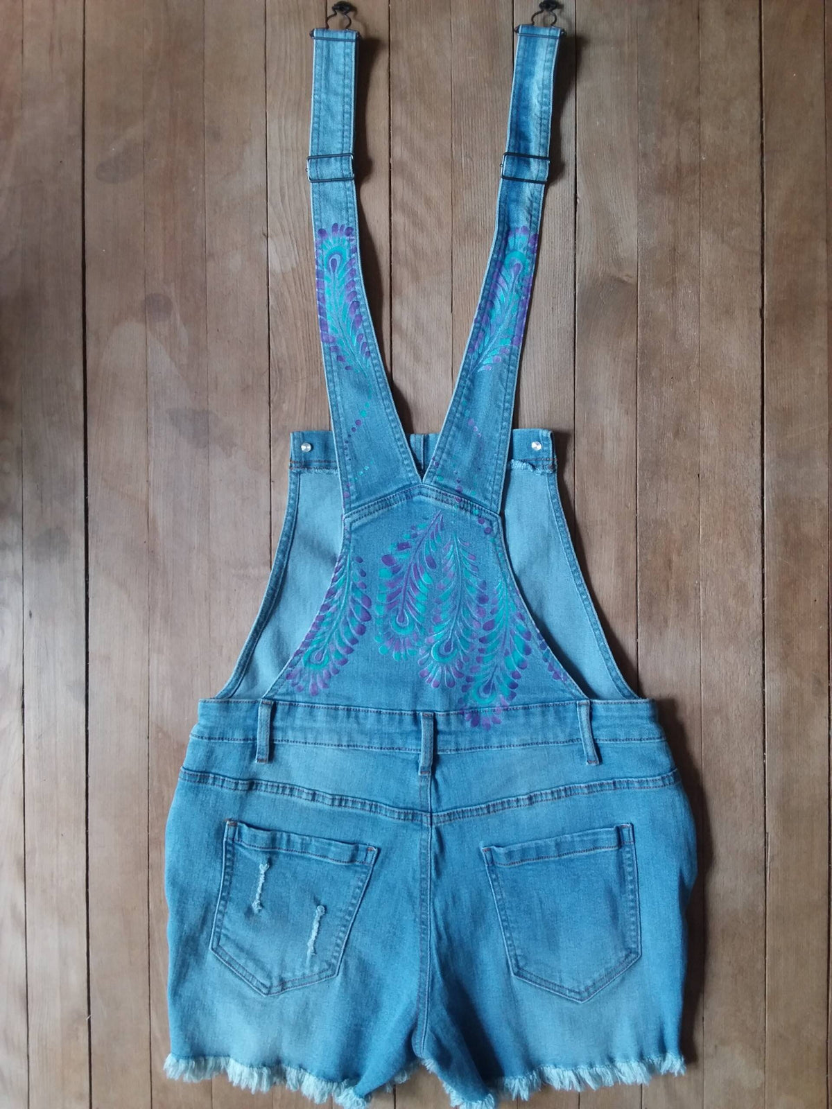 Hand painted ladies denim overalls with distressed details have a lovely purple and teal feather design.
