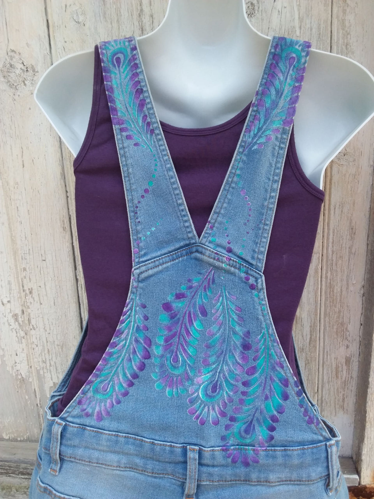 Hand painted ladies denim overalls with distressed details have a lovely purple and teal feather design.
