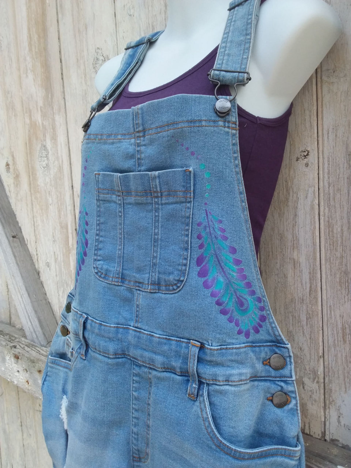 Hand painted ladies denim overalls with distressed details have a lovely purple and teal feather design.