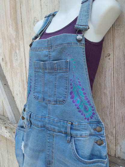 Hand painted ladies denim overalls with distressed details have a lovely purple and teal feather design.