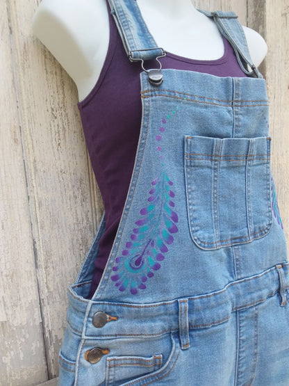Hand painted ladies denim overalls with distressed details have a lovely purple and teal feather design.