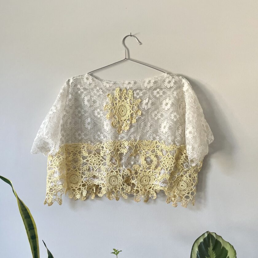 A Boxy Cream Lace Top w/ Crochet Trim hanging on a wall next to a potted plant.