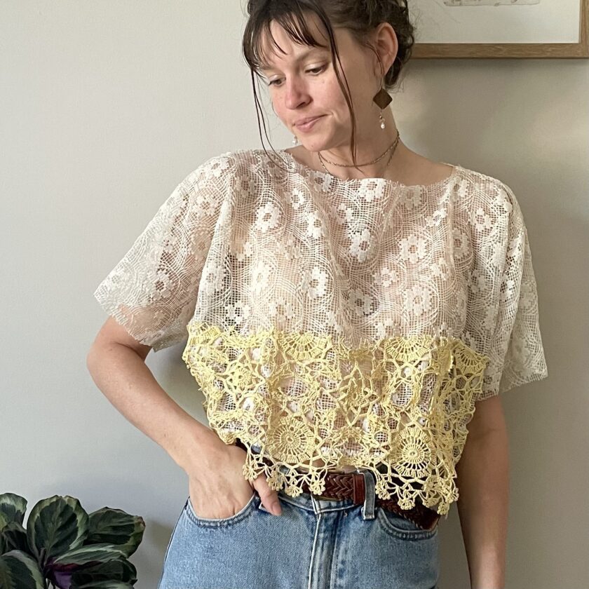 A woman standing in front of a white wall wearing the Boxy Cream Lace Top w/ Crochet Trim.