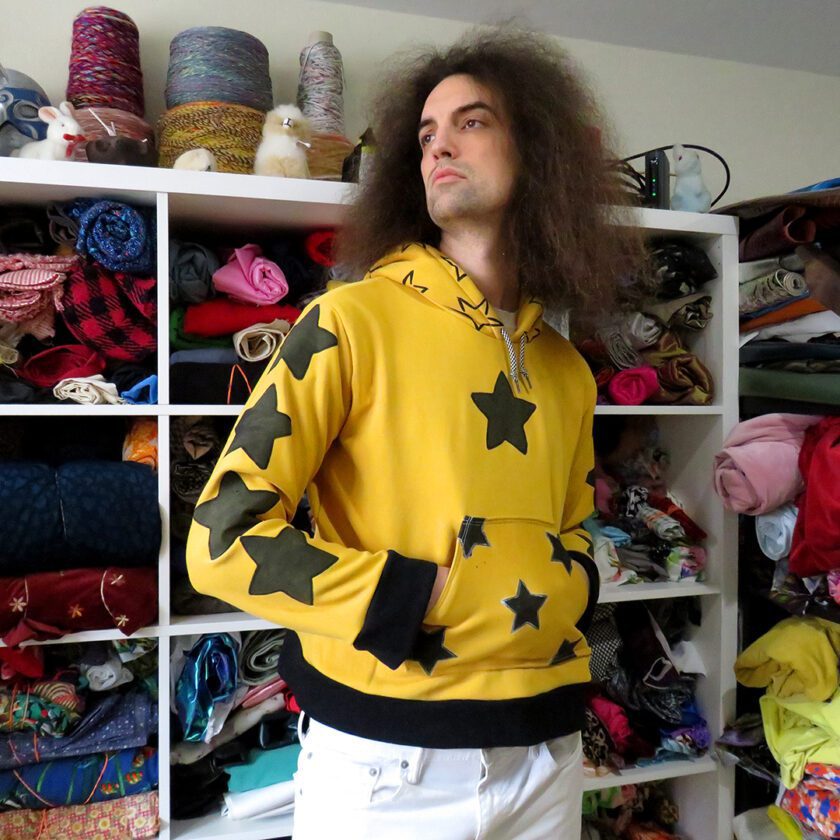 A man with long hair standing in front of a closet full of Yellow Star Hoodies (Large).
