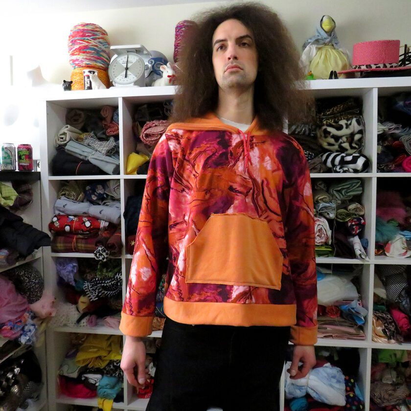 a man with long hair standing in front of a Marbled Orange Hoodie (X-Large) hanging in a closet full of shoes.