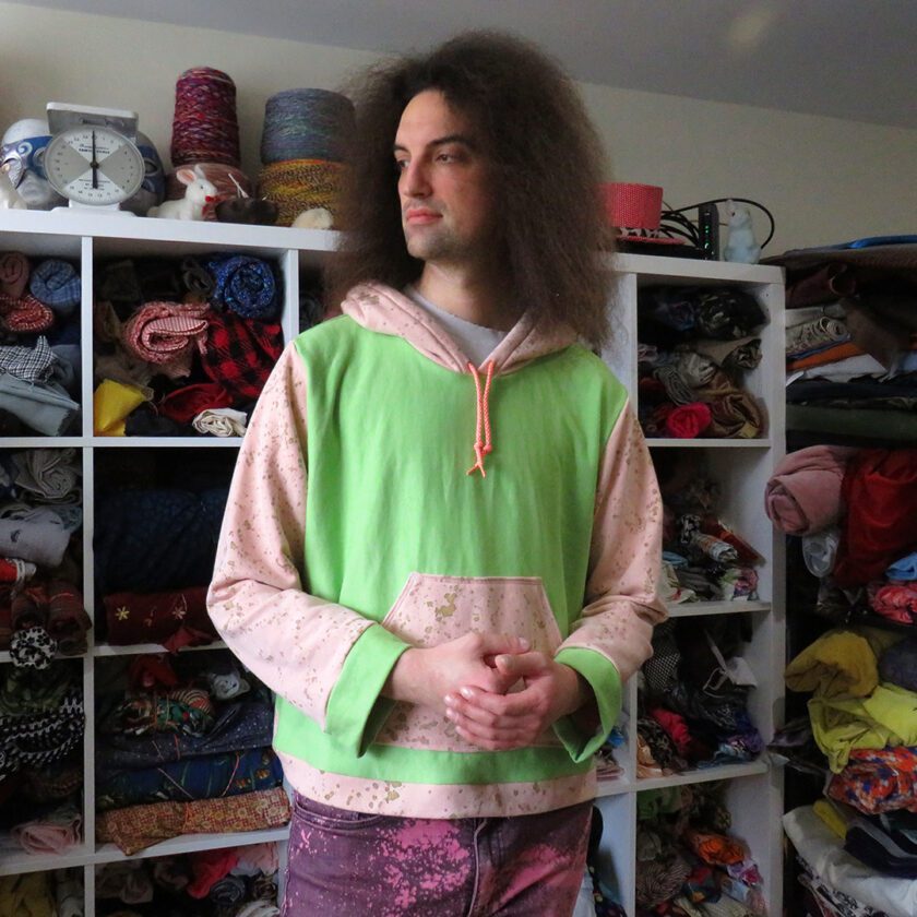 A man with long hair standing in front of a Green and Rusty Pink Hoodie (2X-Large) in the closet.