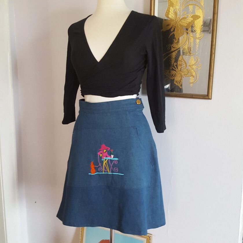 A mannequin wearing an Embroidered Dark Wash Kangaroo Pocket Mini Skirt with a picture of a beach scene on it.