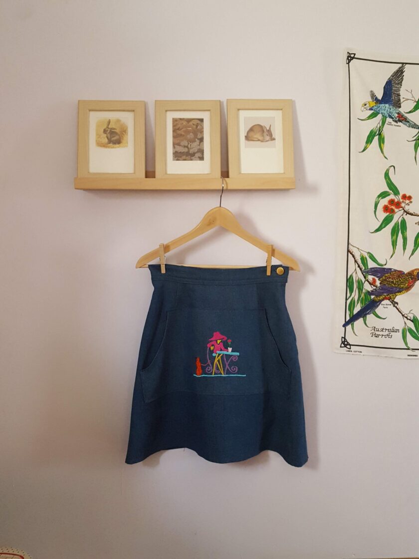 An Embroidered Dark Wash Kangaroo Pocket Mini Skirt hanging up against a wall.