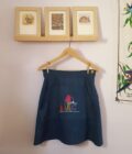 An Embroidered Dark Wash Kangaroo Pocket Mini Skirt hanging on a wall next to two framed pictures.