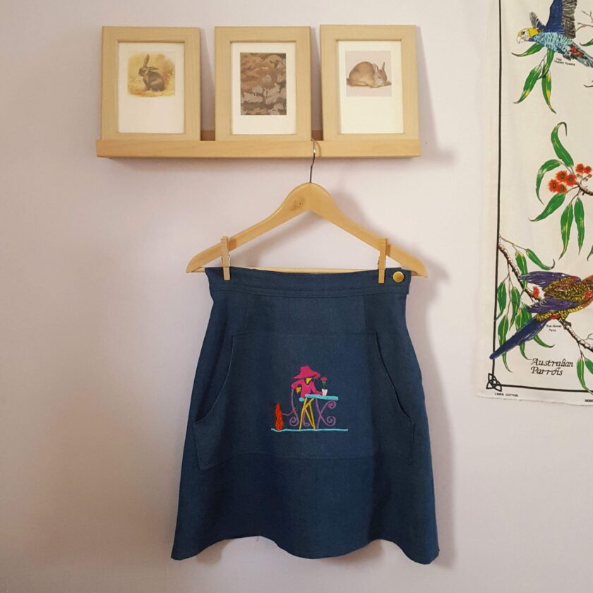 An Embroidered Dark Wash Kangaroo Pocket Mini Skirt hanging on a wall next to two framed pictures.
