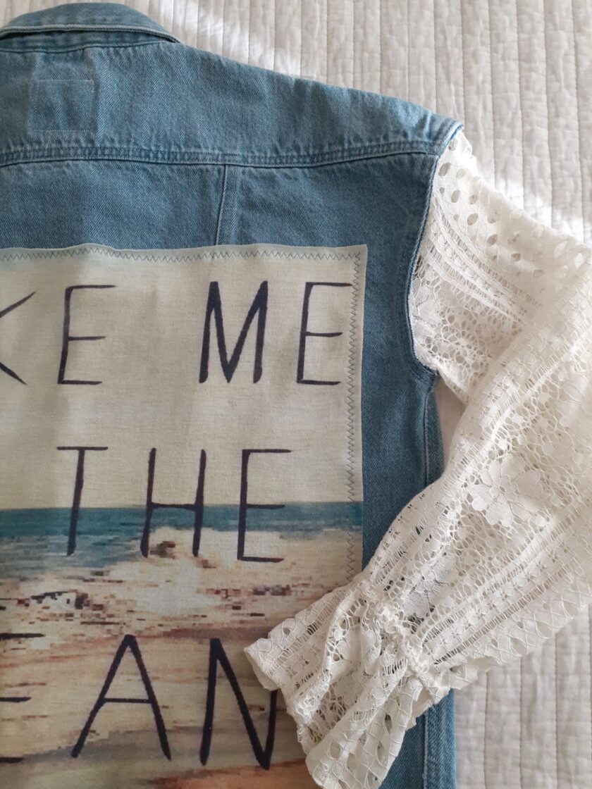 A Take Me To The Ocean upcycled denim & lace jacket with the words take me to the ocean written on it.