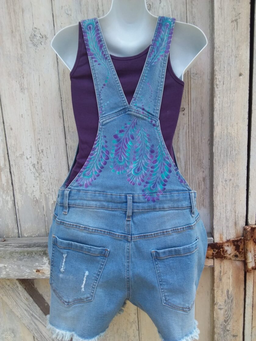 Hand painted ladies denim overalls with distressed details have a lovely purple and teal feather design.