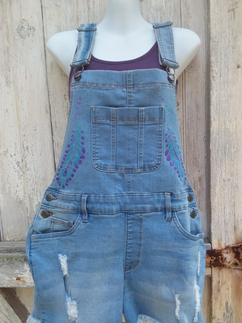 Hand painted ladies denim overalls with distressed details have a lovely purple and teal feather design.