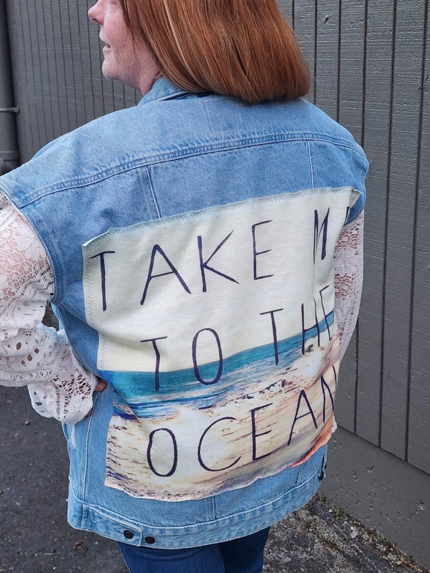 A woman wearing a Take Me To The Ocean upcycled denim & lace jacket with a message painted on it.
