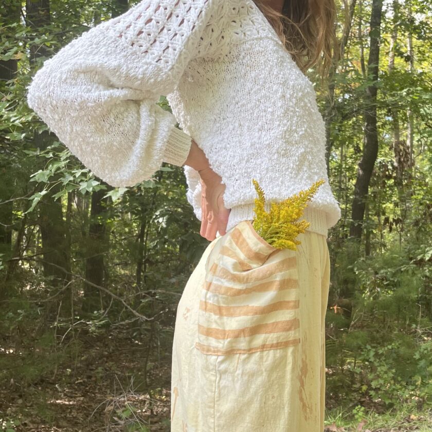 A woman is standing in the woods wearing a sweater and skirt.