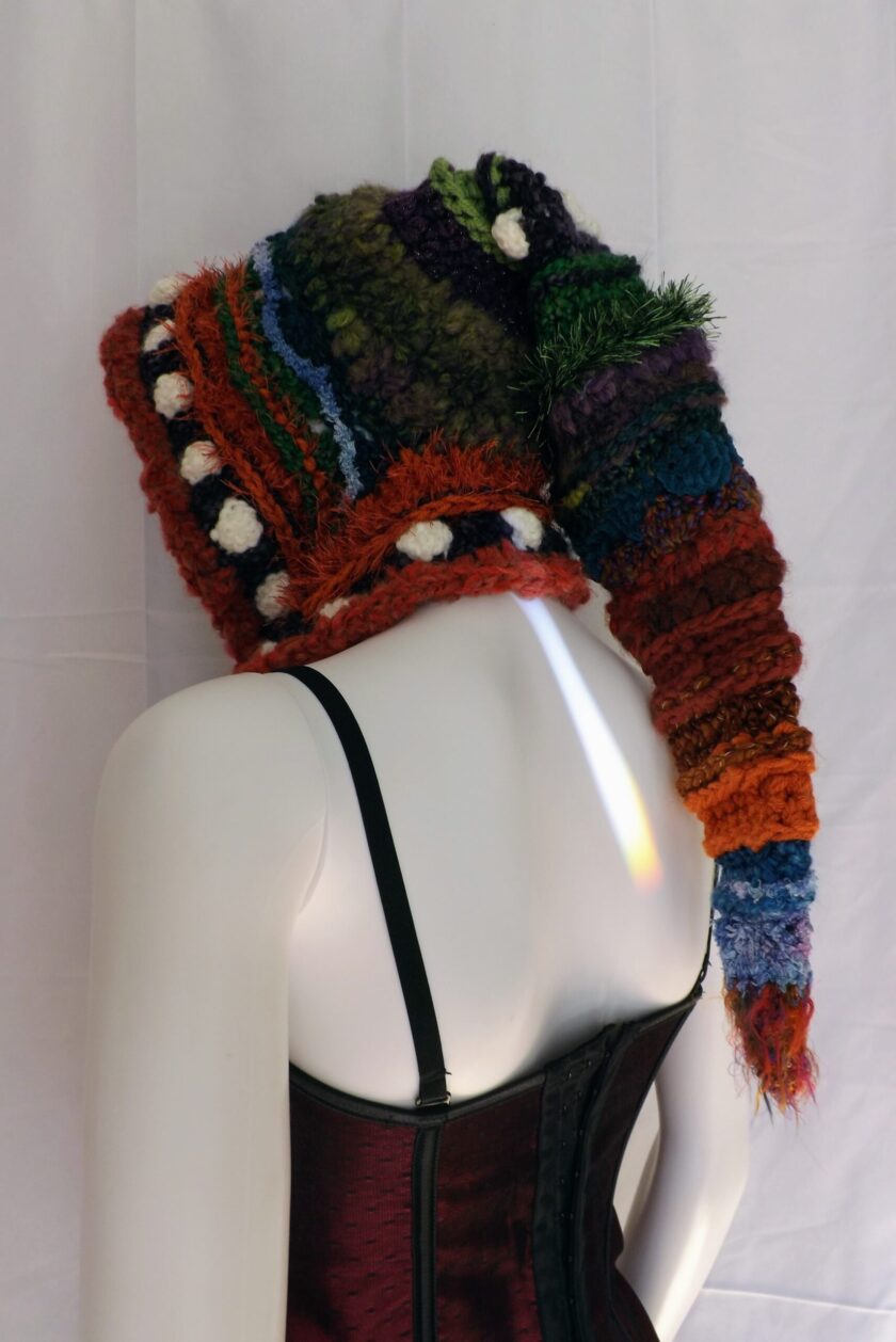 A crochet elf hood in greens, purples, orange and blue on a mannequin