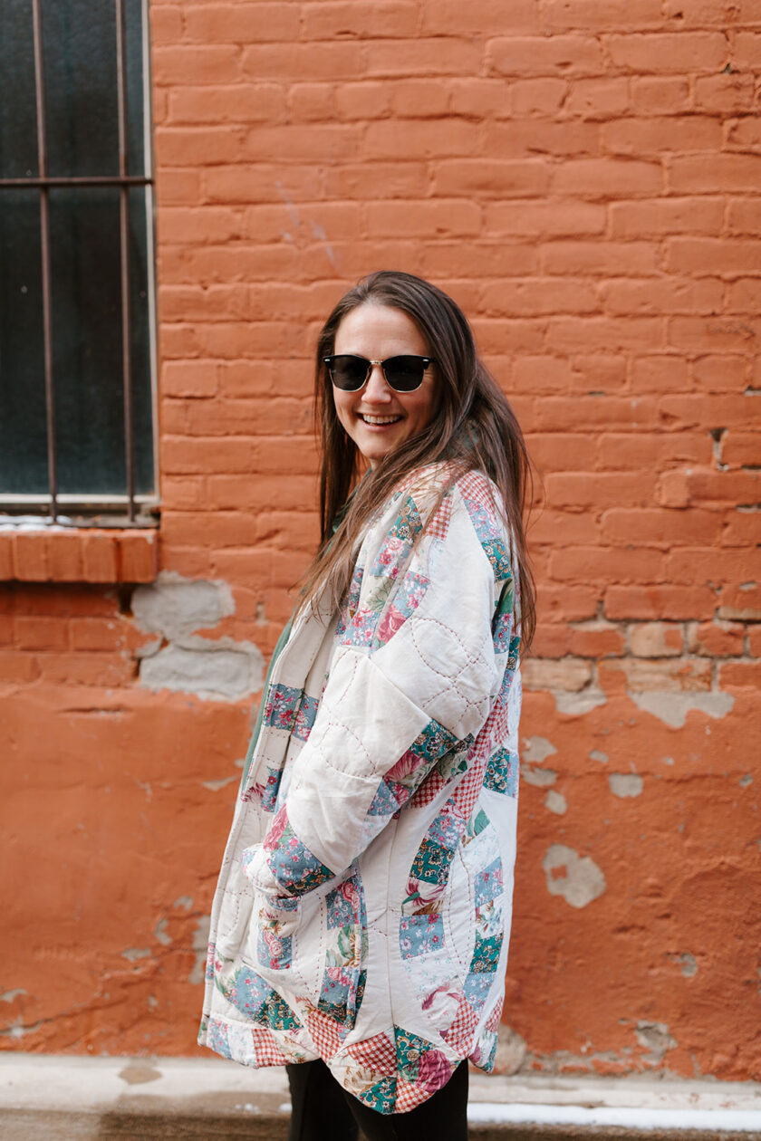 A woman wearing a colorful quilted jacket in front of a brick wall.