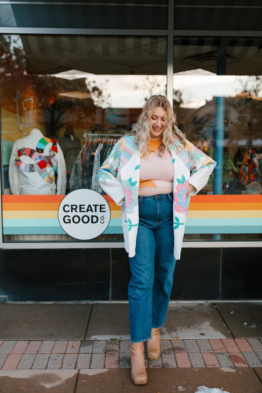 A plus size woman in jeans and a colorful cardigan standing in front of a store.