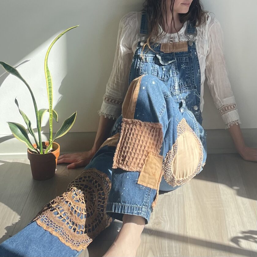 A woman in overalls sitting on the floor next to a potted plant.