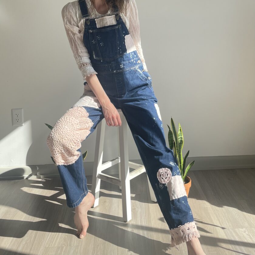 A woman in overalls sitting on a stool.