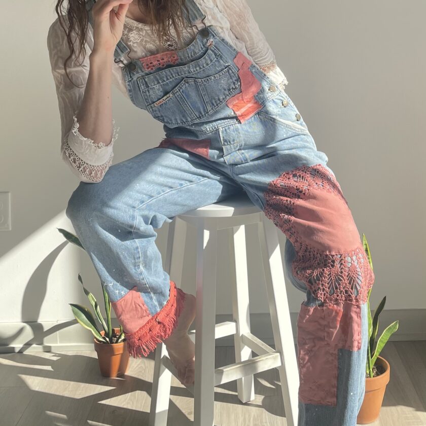 A woman in overalls sitting on a stool.
