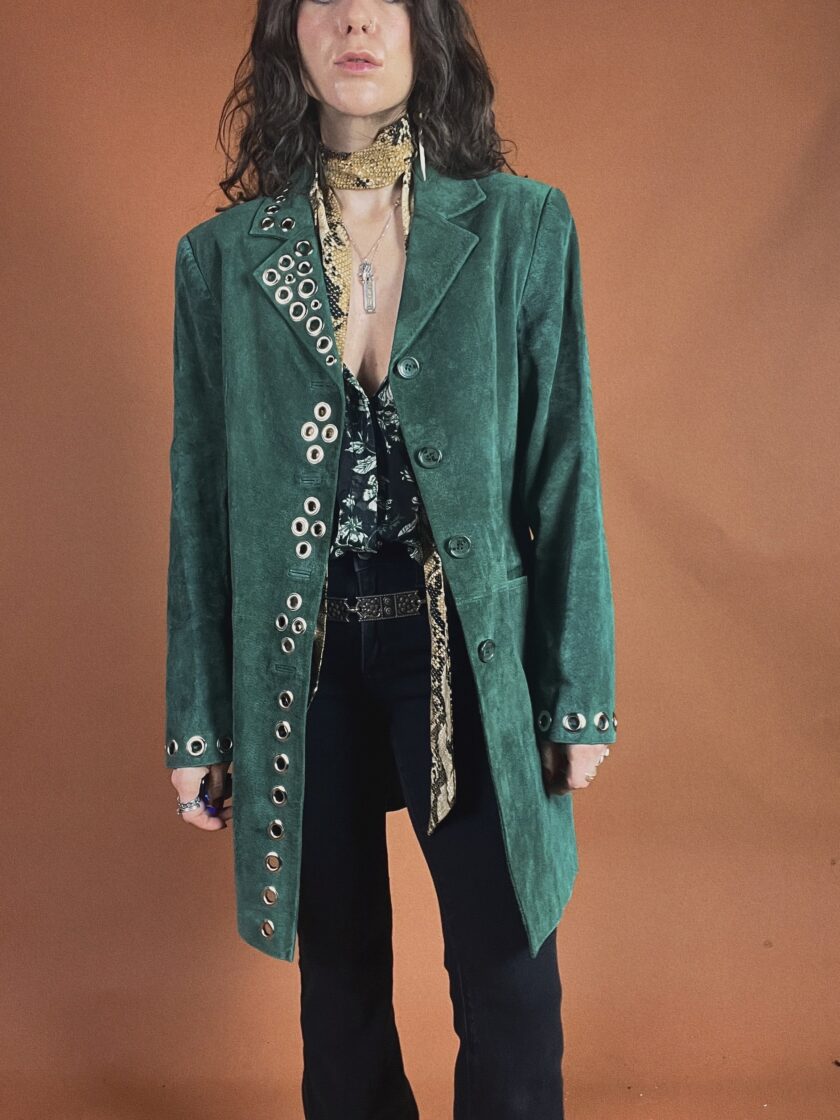 Woman posing in a green velvet coat with metallic button details, styled with a patterned scarf and black trousers.