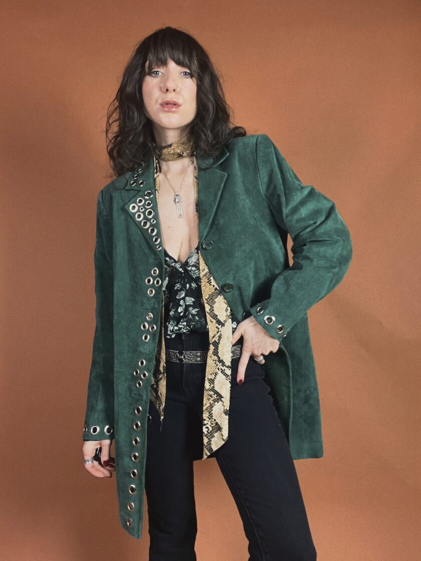 Woman posing in a stylish green jacket and black pants with a patterned blouse and accessories.