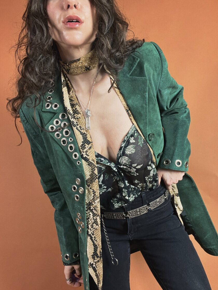 Woman posing in a green velvet blazer with decorative buttons, patterned scarf, and black pants.