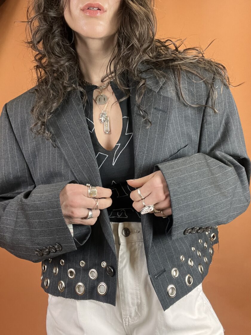 Woman wearing a pinstripe blazer with grommet details, paired with a black top and white trousers, accessorized with multiple rings and a pendant necklace.