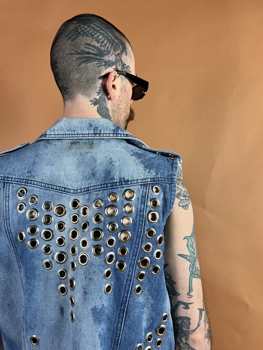 Man with a tattooed scalp and arm wearing a studded denim vest and sunglasses, viewed from behind against a brown background.