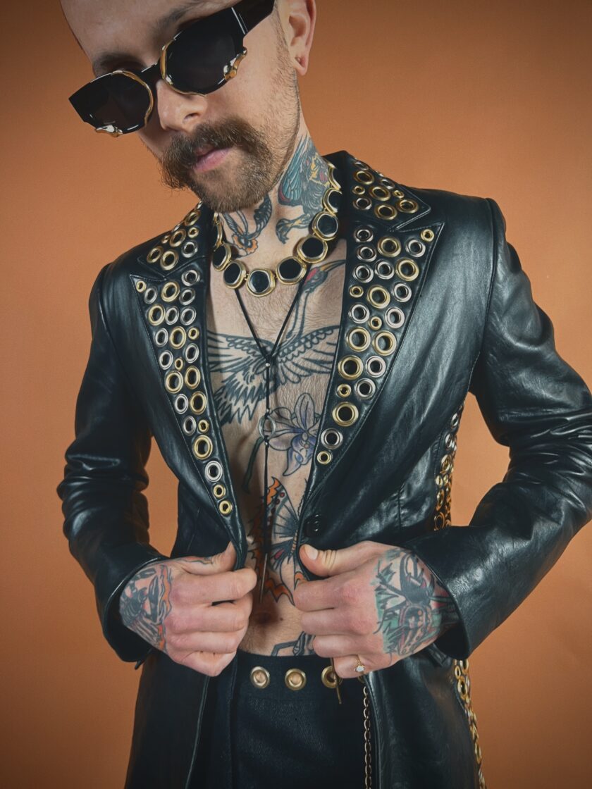 A man with a beard and tattoos wearing sunglasses and a black leather jacket with circular cut-outs.
