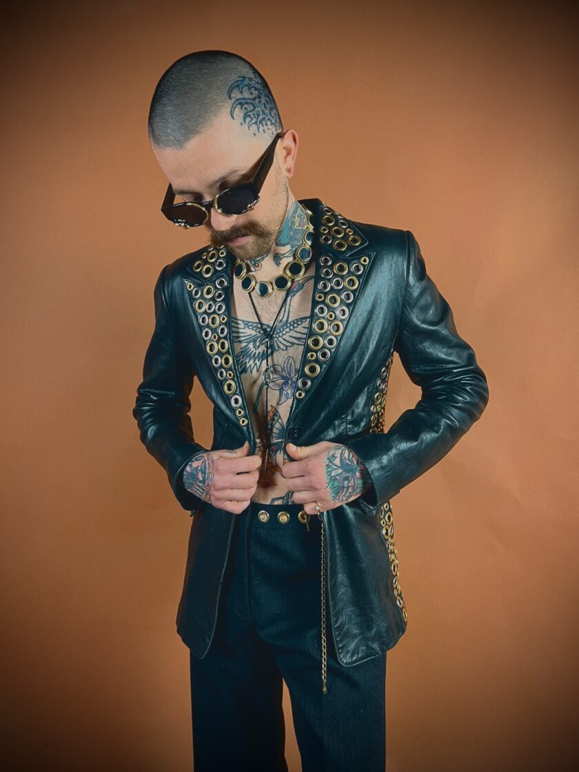 A person with a shaved head and tattoos stands wearing sunglasses, a leather jacket, and ornate clothing.